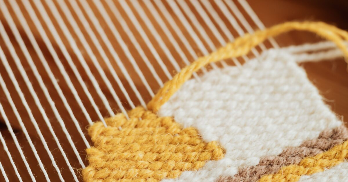 Close-up of a hand loom: taut warp threads on the left flowing into a half-finished woven panel in mustard, cream, and tan — a pattern being built thread by thread.